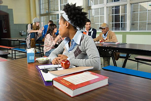 A student sitting alone doing homework and eating. This student does this while watching the group of students behind her. This group is seen as being social with one another leaving the student sitting at an empty table by herself. Source: Getty Images