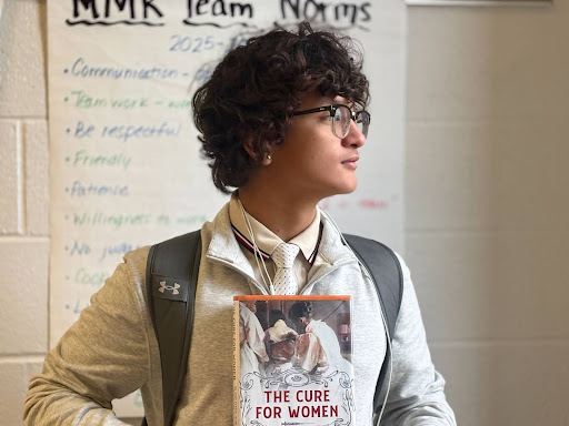 Sophomore Aiden Bell, posing for the camera by wearing a quarter-zip, holding up his feminist literature and looking into the distance. 